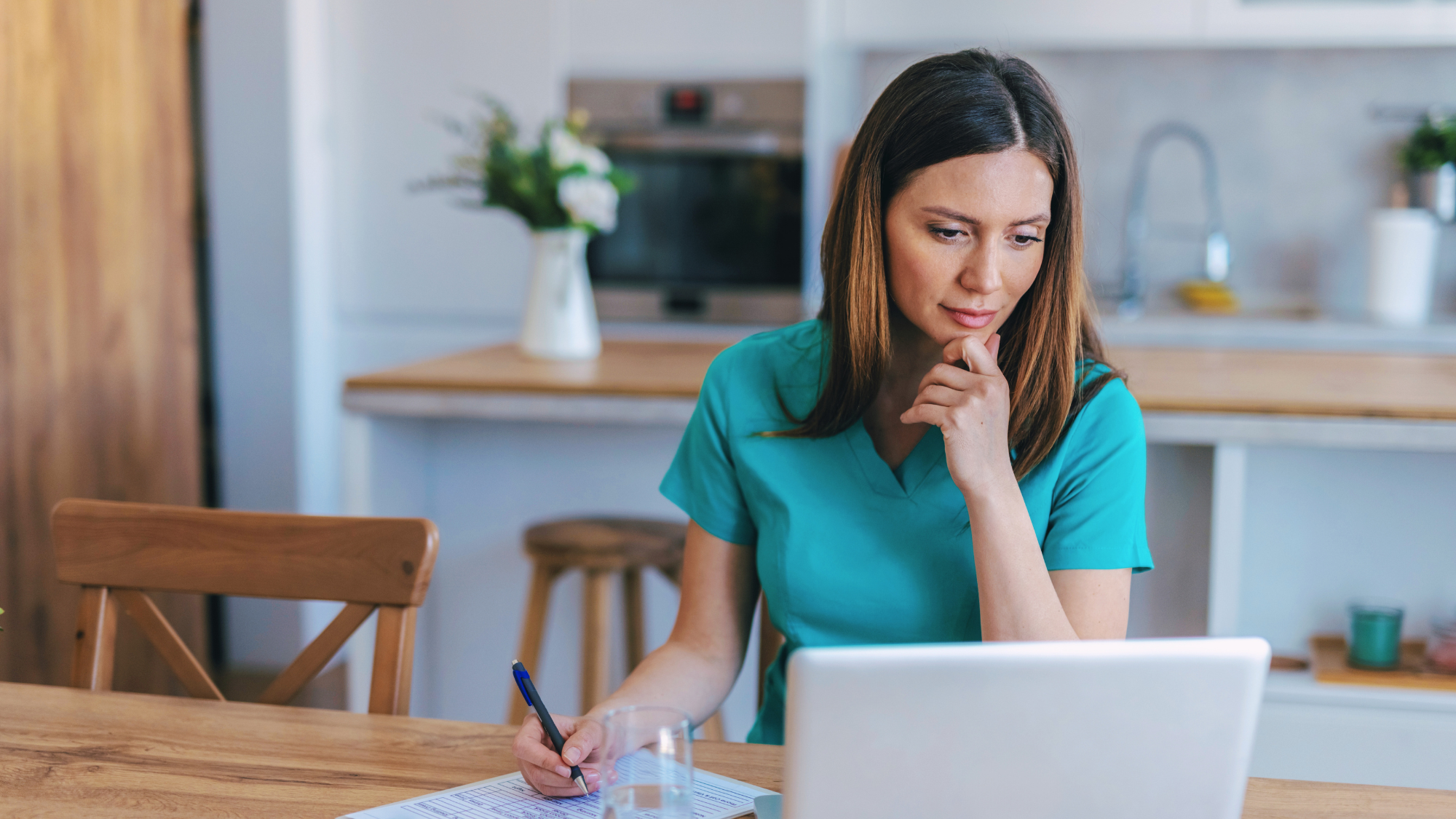 Female nurse in scrubs sitting at a desk in her home looking at a computer.