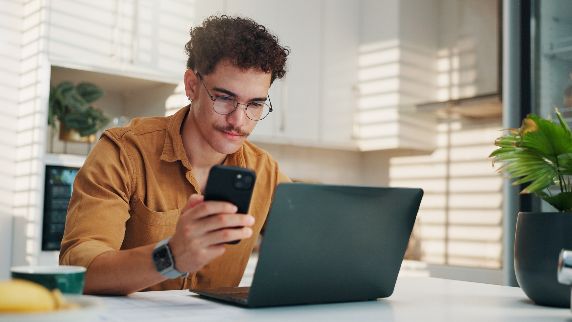 Man with brown hair and glasses looking at his laptop and phone in a kitchen.