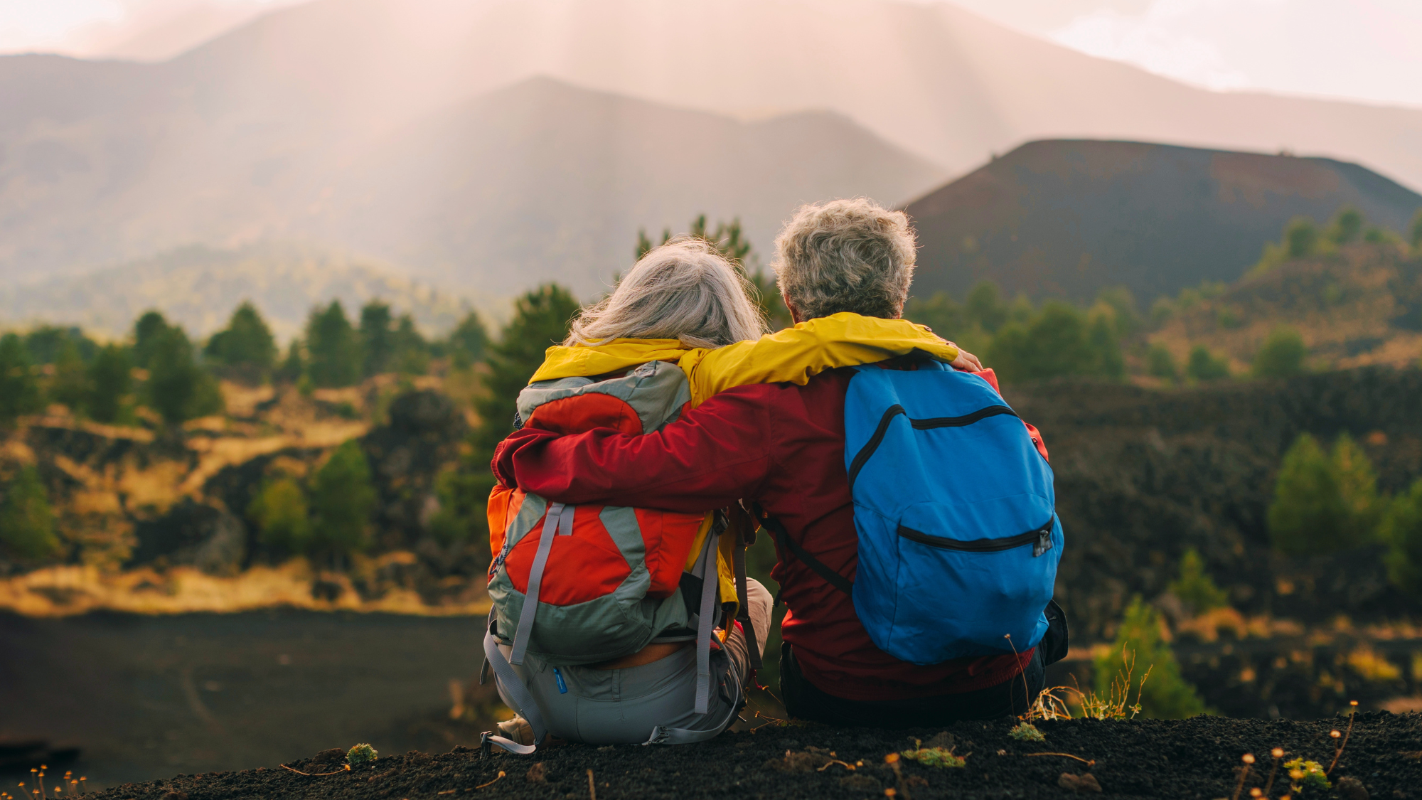 Two elderly individuals sitting on ledge wearing backpacks with the arms wrapped around each others shoulders.